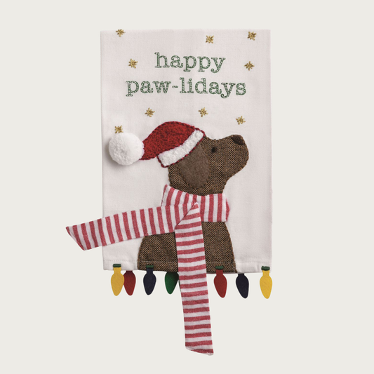 Christmas-themed white decorative towel, with a brown labrador-type dog wearing a Santa hat and red & white scarf that trails off the edges of the towel. Text above it reads: "Happy Paw-Lidays". Small gold stars are floating around the text. Felt holiday light bulbs are hanging from the bottom of the towel.