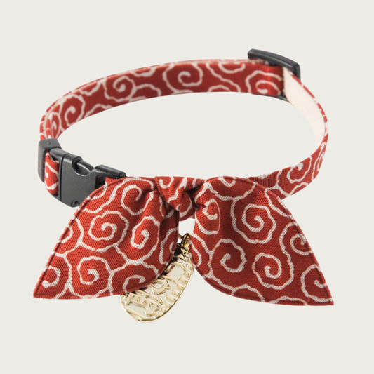 Cat collar with a deep rust-colored background and white curly-q pattern, with a simple bow in the same pattern. It has a dangling gold tone Asian-inspired charm and black plastic clip & slider.