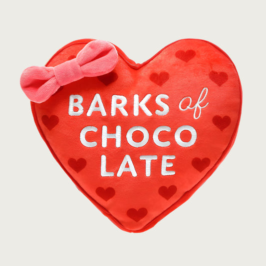 Valentine's themed, plush dog toy that looks like a heart-shaped red box of chocolate. Centered, large, white text reads 'BARKS of CHOCOLATE'  and there's a pink bow on the top left of the toy.