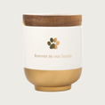 Load image into Gallery viewer, Cream-colored ceramic pet memorial jar with dark brown wooden lid. Bottom 1/3 of jar has a matte gold finish. Minimalist design in center features a gold paw print, with "forever in our hearts" text in gold underneath it.
