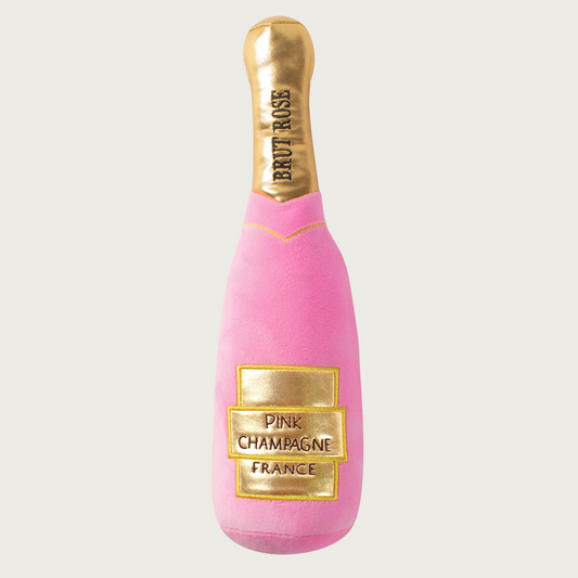Pink plush dog toy shaped like a champagne bottle with gold label on body & neck.