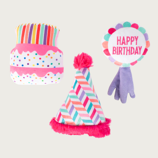 Three mini birthday themed plush dog toys. Cake looks like a 2-layer white cake with multicolored sprinkles and pink drip frosting, with "candles" on top. Birthday Hat has an hot pink rim & pom pom at the top. Its body features a multicolored chevron design. The Ribbon is shaped like a stereotypical blue ribbon award, with 2 lavender tails, and round white body that says "Happy Birthday" in hot pink text, bordered with a multicolored scalloped design edge. 