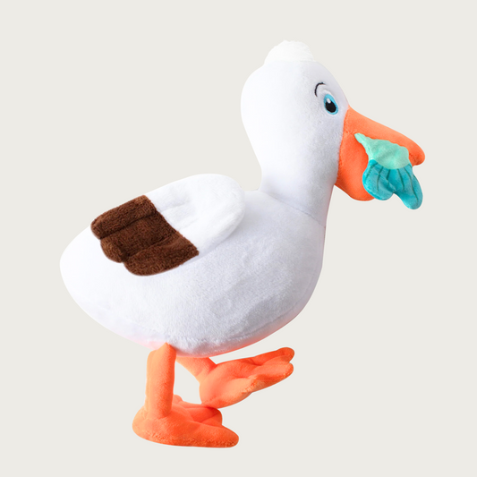 Plush seagull dog toy with white body, brown tipped wings, and orange beak and feet. It has a teal blue fish tail sticking out of its beak.