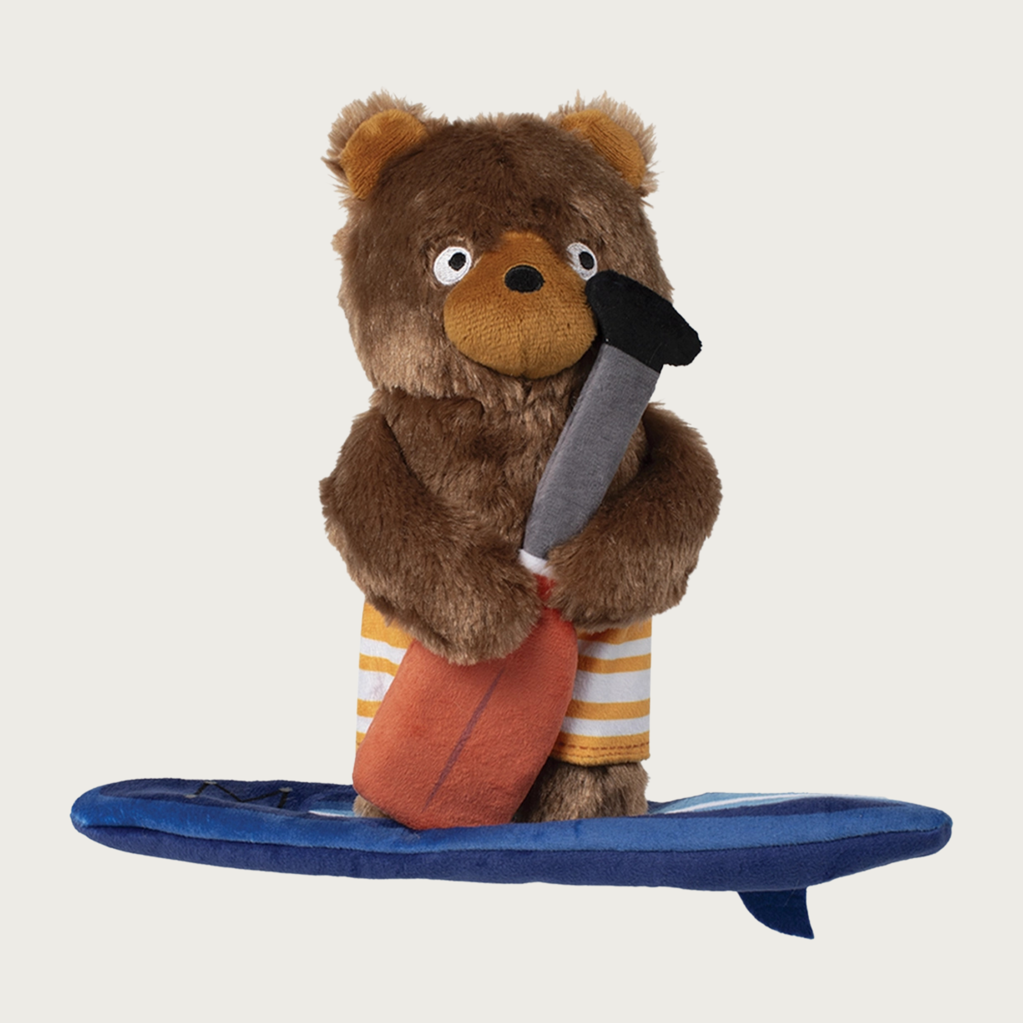 Plush brown bear dog toy standing on a plush blue paddleboard, and holding a long paddle. The bear is wearing orange & white striped board shorts.