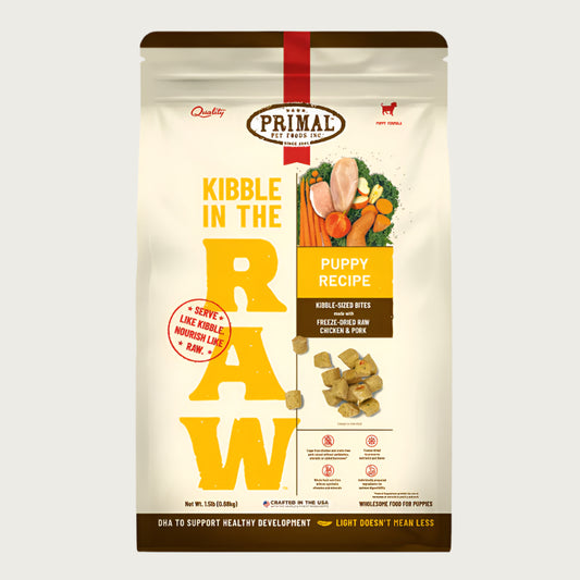 A 1.5 pound bag of puppy recipe Kibble in the Raw dog food from Primal Pet Foods. Text on bag reads "Kibble sized bites made with freeze-dried raw chicken & pork" and "Serve like kibble. Nourish like raw." There is an image of fresh produce with a raw chicken breast and pork chop, and a smaller image of the bite-sized morsels that are in the bag.
