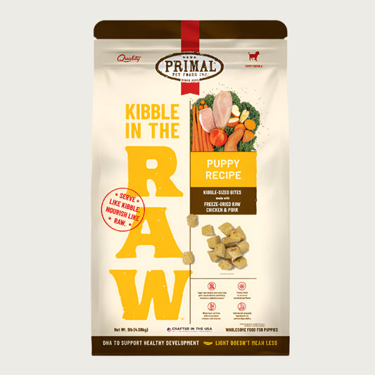 A 9 pound bag of puppy recipe Kibble in the Raw dog food from Primal Pet Foods. Text on bag reads "Kibble sized bites made with freeze-dried raw chicken & pork" and "Serve like kibble. Nourish like raw." There is an image of fresh produce with a raw chicken breast and pork chop, and a smaller image of the bite-sized morsels that are in the bag.