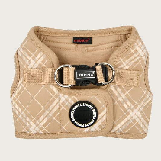 View of Puppia Corwin Step-In vest-style harness for dogs, laid flat. Interior and trim is tan colored, with the exterior cream plaid/check print on tan. Fastener clip on back is black. Accented with black rubber circle behind it, that says Puppia Sports in a repeating circular pattern.