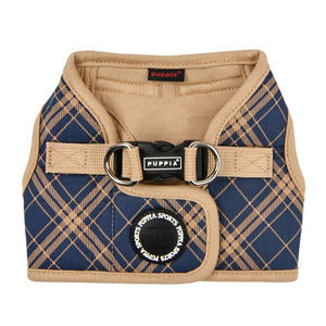 View of Puppia Corwin Step-In vest-style harness for dogs, laid flat. Interior and trim is tan colored, with the exterior tan plaid/check print on navy blue. Fastener clip on back is black. Accented with black rubber circle behind it, that says Puppia Sports in a repeating circular pattern.