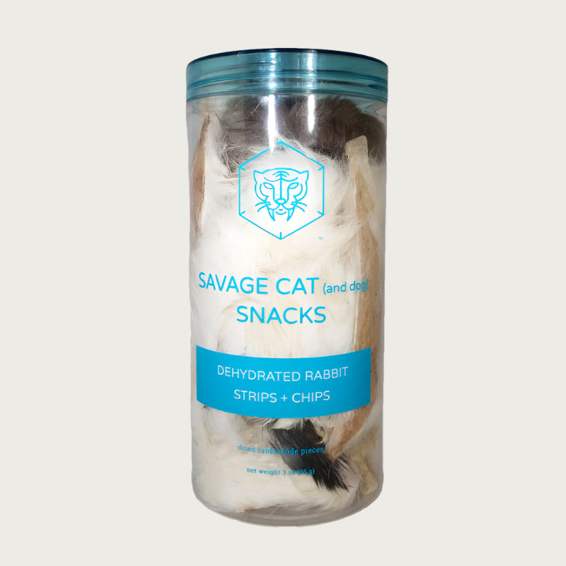 A 3 oz clear plastic container, filled with dehydrated rabbit strips and chips (rabbit hide pieces) by Savage Cat. Fur covered pieces in various natural shades from brown to white are visible.