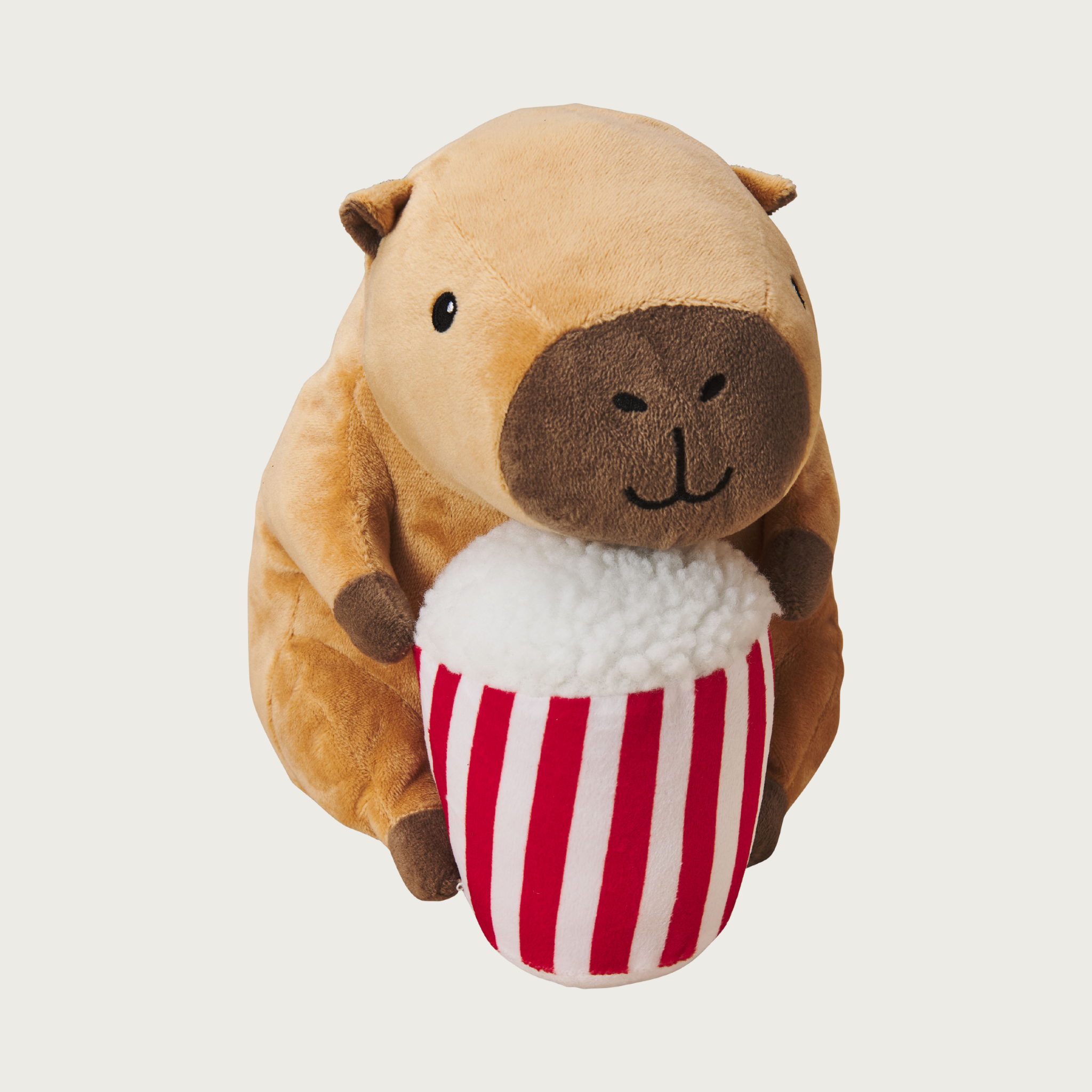Plush, plump, brown capybara dog toy holding a red & white striped plush popcorn tub in its hands. Has a cute embroidered facial expression.