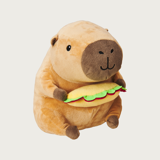 Plush, plump, brown capybara dog toy holding a plush taco in its hands. Has a cute embroidered facial expression.