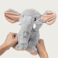 Load image into Gallery viewer, A plush gray elephant dog toy being squeezed by two hands (one on each side of its body). The elephant has white tusks, pale pink inner ears and underside of its feet, and embroidered black & white eyes.
