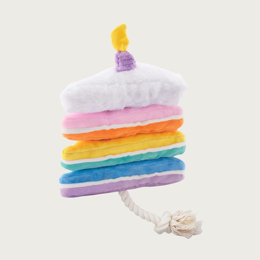 Triangle shaped dog toy designed to look like a slice of layered birthday cake. There's multiple pastel hued layers, with chunky white (frosting) layer on top, with a corduroy birthday candle. All are connected with a cream colored rope.
