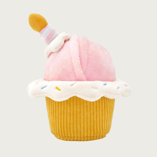 Cupcake-shaped plush dog toy. Round top is pale pink, base is deep yellow corduroy plush, with a "frosting" accent in-between that's white with multicolored embroidered pastel sprinkles. Pointing out of the pink top is a plush birthday candle.