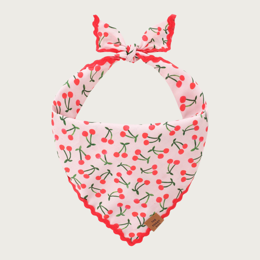 Dog bandana with a blush pink background, featuring a pattern of pairs of red cherries with green stems & leaves, and red scalloped edging.