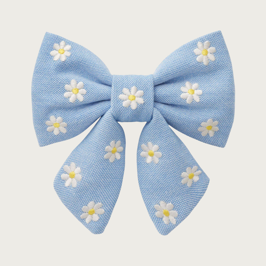 Light blue Oxford cloth lady bow tie for dogs, with embroidered white daisies with yellow centers, stitched evenly across the entire bow. Looks like a hair bow for a person, with the center bow tie and ends hanging down.