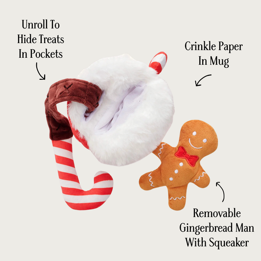 View from the top of the cocoa mug toy, showing two openings. The candy cane toy is out of the mug, but attached to it with a wide brown plush ribbon. It is labeled with "unroll to hide treats in pockets" text. The gingerbread man toy is pulled out and labeled with "removable gingerbread man with squeaker" text. 