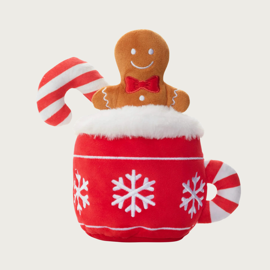 Plush dog toy in the shape of a cocoa mug, with a toy candy cane and gingerbread man sticking out of the top of the mug. The mug is red with white embroidered snowflakes and a candy cane striped handle, and fuzzy faux white fur on the rim.