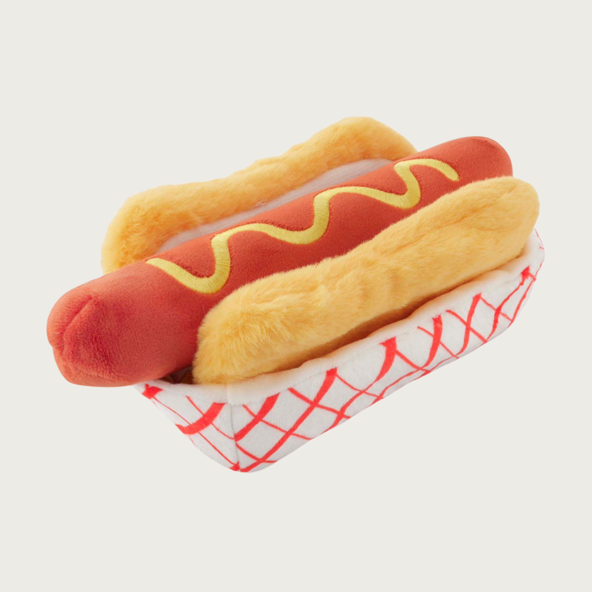 Plush dog toy designed to resemble a hot dog in a paper container. It has a light yellow bun, reddish-pink hot dog with mustard drizzle, set in a white "paper" container with red diagonal stripes.