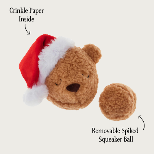 Light brown plush teddy bear head shaped dog toy wearing a red Santa hat, with removable spiked squeaker ball next to it. Ball has the same light brown plush covering it. Text pointing to the teddy bear says "crinkle paper inside" and text pointing to the smaller ball says "removable spiked squeaker ball".