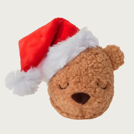 Light brown plush teddy bear head shaped dog toy wearing a red Santa hat. Simple, sleepy, embroidered facial expression. 