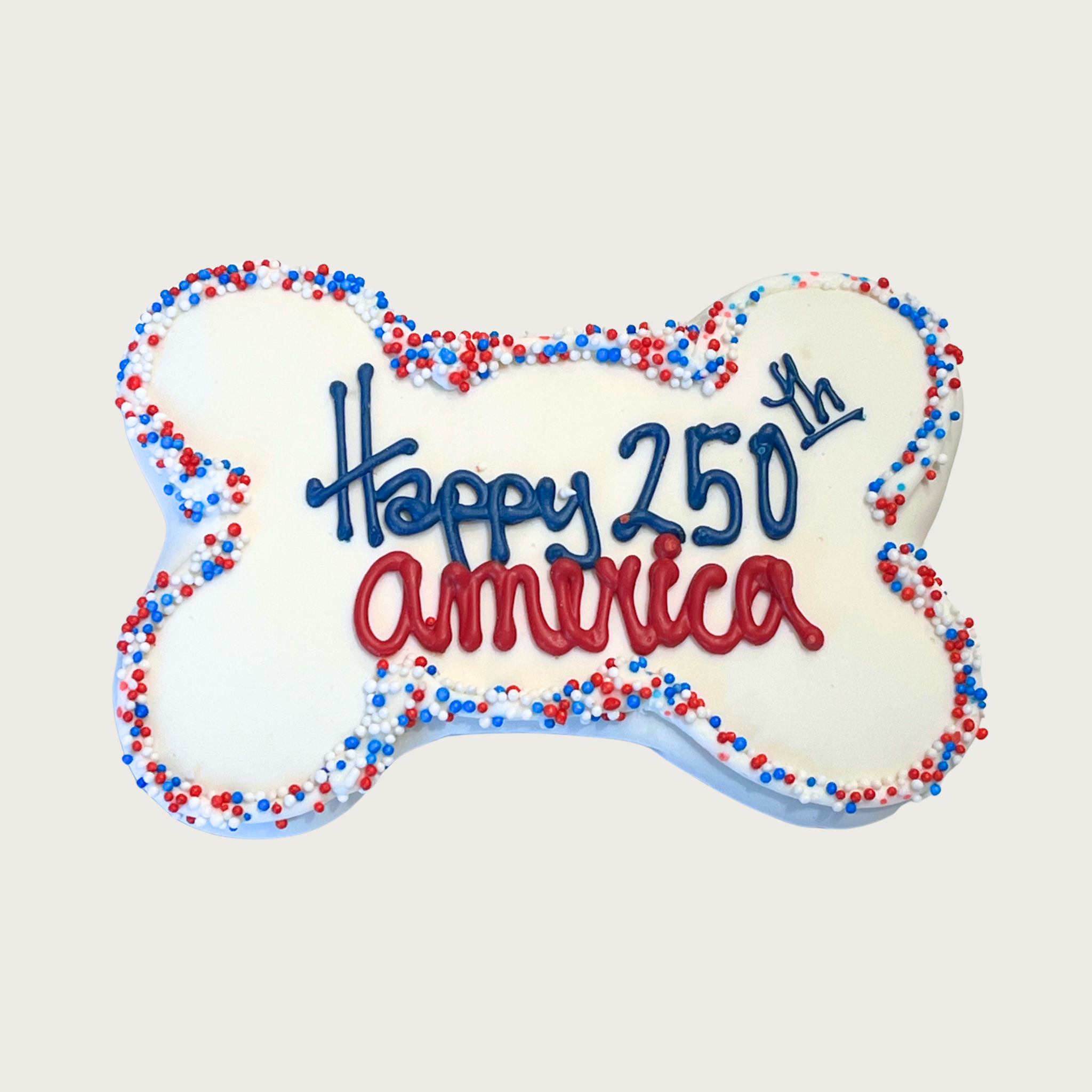 Dog bone-shaped dog cookie with 'Happy 250th America' text in center. "Happy 250th" is in navy blue, and "america" is in red. A thin border of red, white, and blue nonpareil sprinkles decorates the edges of the cookie.