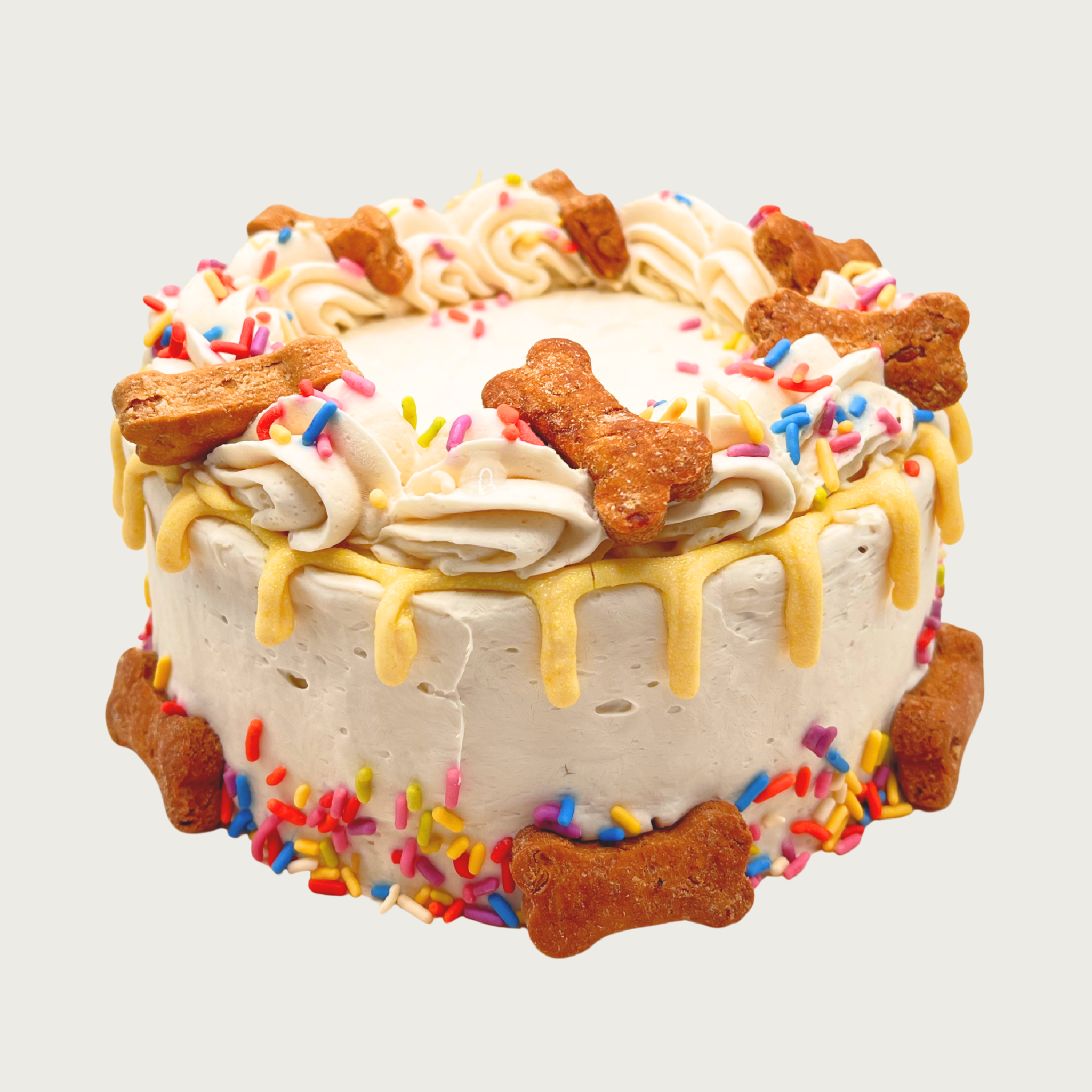 A cream colored dog birthday cake with piped frosting, yellow drip accent, multicolored sprinkles, and accented with bone-shaped dog biscuits.