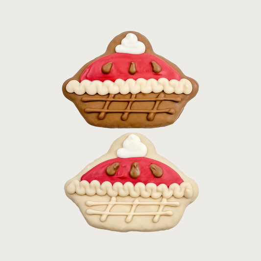 Two decorative dog cookies shaped like whole pies. One is primarily dark brown with red filling & whipped cream. The other is primarily light brown with the same decoration.