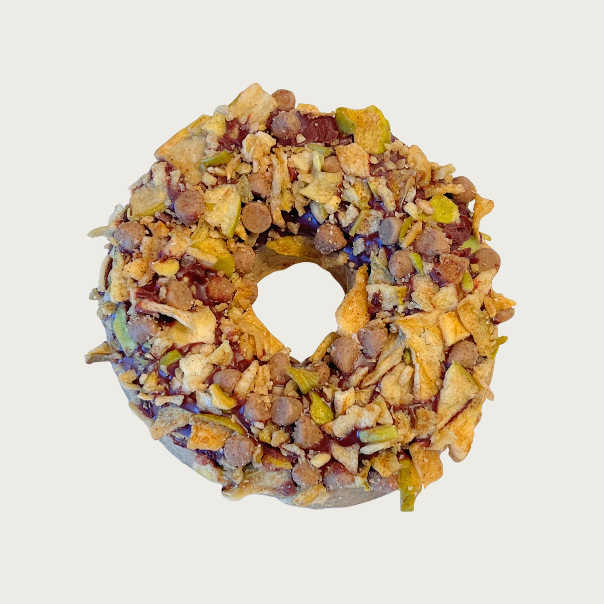 A thick, donut shaped dog cookie, covered with dried apple pieces, tan mini cinnamon chips, and granola bits.