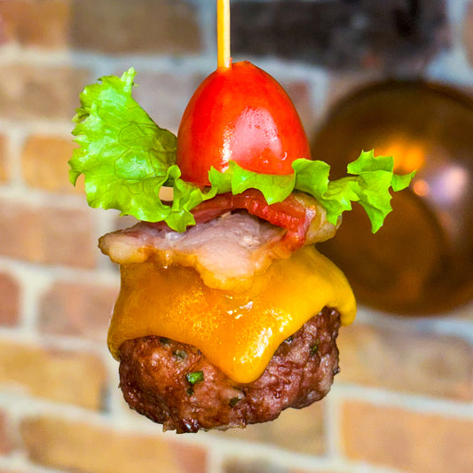 Extreme close-up of a bacon cheeseburger bite dog treat on a skewer. The base is a meatball with cheddar cheese melted over it, with a small piece of bacon, and lettuce and half a grape/cherry tomato on top.