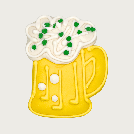 Dog cookie shaped like a yellow beer mug with white foam and green clover shaped sprinkles.