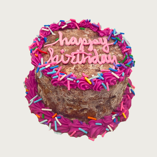 Small birthday cake for dogs. It looks like a rounded frozen meatloaf accented with deep pink frosting and colorful sprinkles around the edges. "Happy Birthday" is written in pink script text in center.