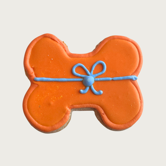 Dog bone-shaped decorated dog cookie with orange icing and an iced light blue bow with ribbon across the center.