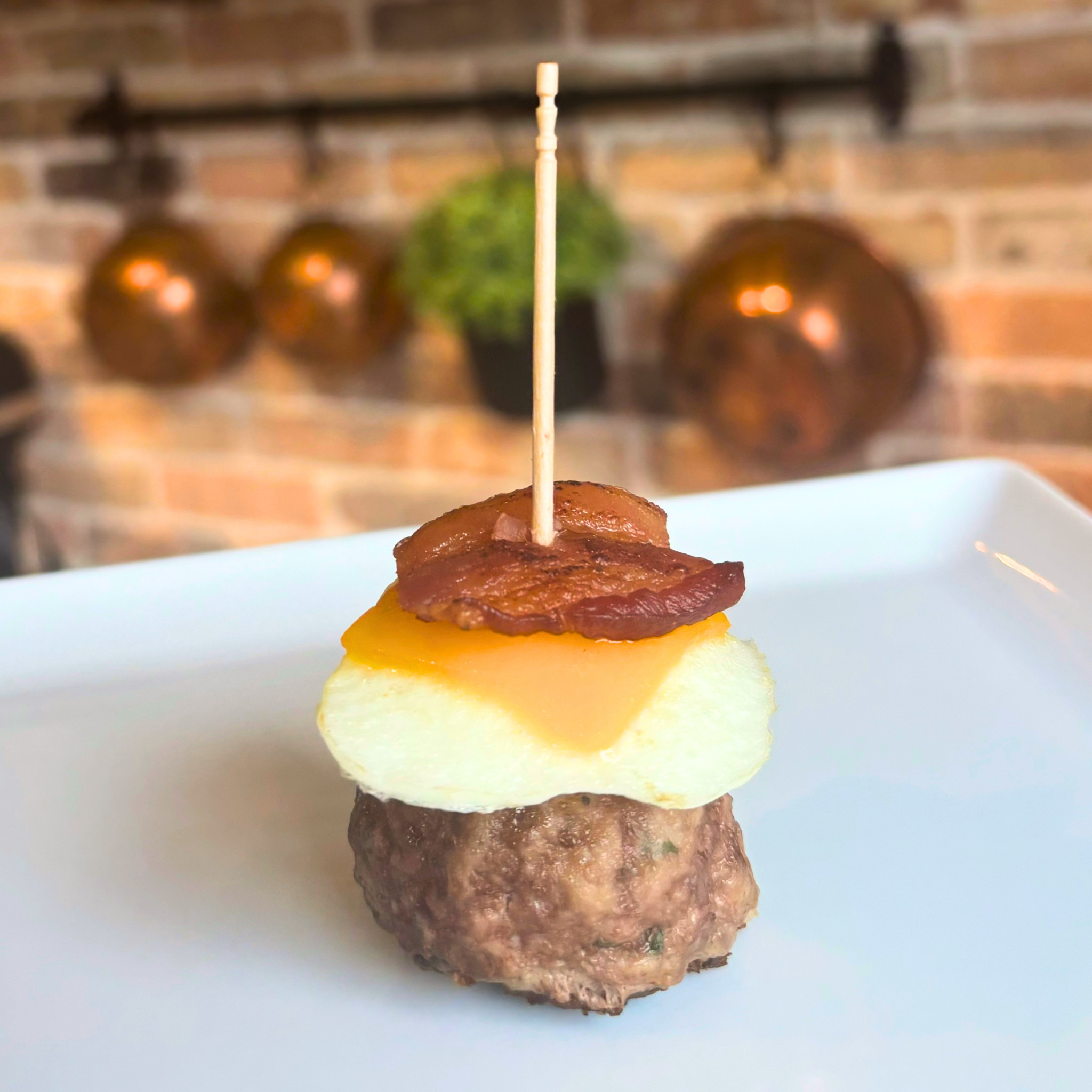 A Breakfast Burger Bite dog treat, starting with a large meatball, topped with fried egg, melted cheddar cheese, and a crispy small piece of bacon, held together by a wooden toothpick on a white plate.