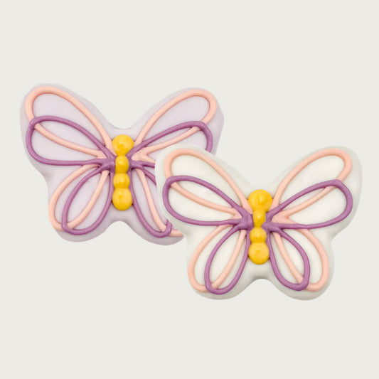 Two butterfly-shaped dog cookies with pink, purple, and yellow icing on a white or pale purple body.