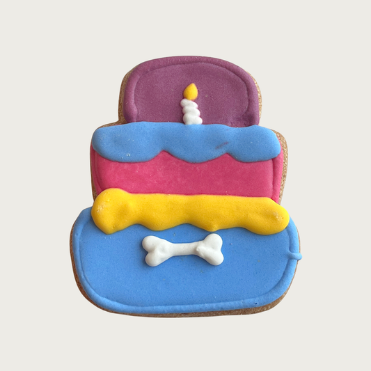 Decorated dog cookie shaped like a three-tiered birthday cake. Bottom tier is iced blue, middle is pink, and top is purple. The frosting "drip" between layers is yellow then blue. Small white candle with flame detail is on top, with a white dog bone centered on the bottom tier.