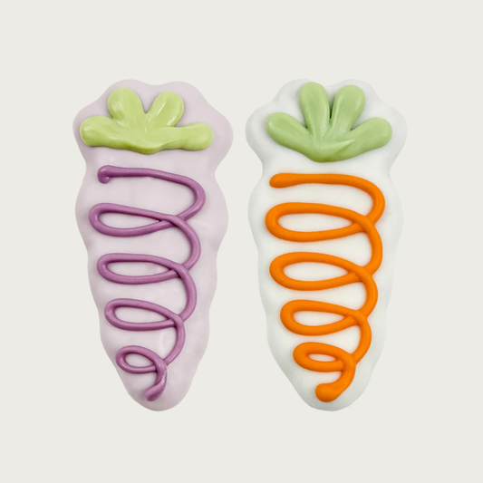 Two carrot-shaped dog cookies side by side. The one on the left is coated in pastel purple frosting, accented with pale green leaves & dark purple squiggle down the body. The one on the right is coated in white frosting, accented with pale green leaves & orange squiggle down the body. 