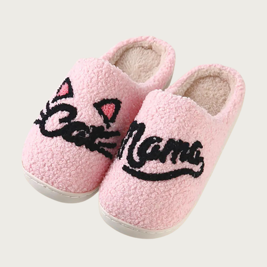 Plush sherpa, pale pink slippers with "Cat" on the right one and "Mama" on the left, written in black script text. The "Cat" slipper has whiskers on both sides of the word, with triangular pink & black cat ears above the word.