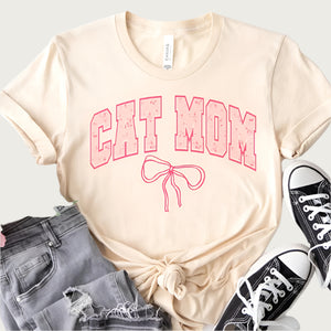 Cream colored short sleeve t-shirt with Cat Mom in large, varsity style lettering on the chest. Border color on text is hot pink, interior text color is pastel pink with tiny floral accents. There is a hot pink bow detail placed underneath the text.