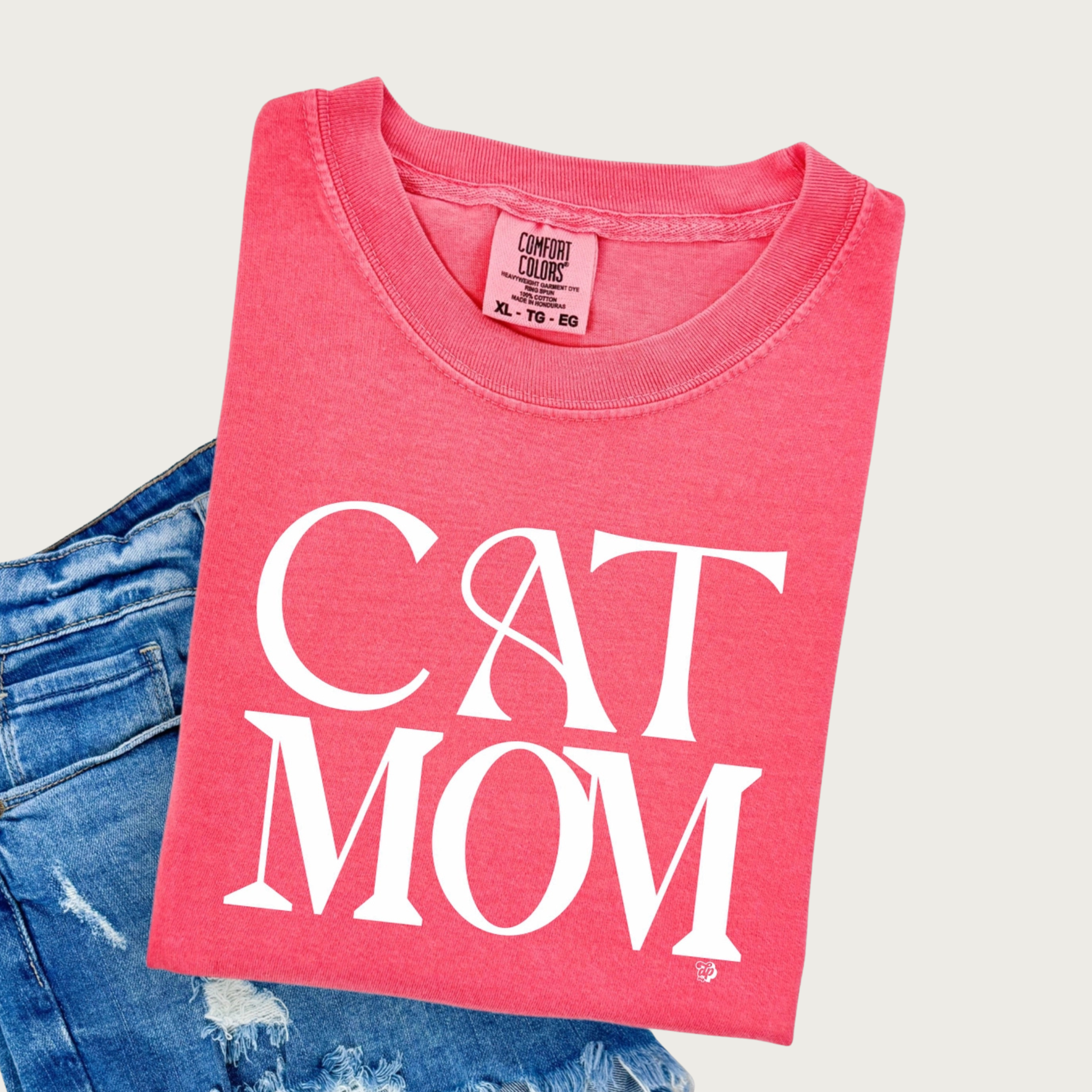 A folded, bright coral pink, crewneck, short sleeved t-shirt with 'CAT MOM' printed in large white art deco style, all caps letters, styled on top of distressed blue denim shorts.