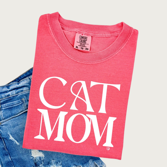 A folded, bright coral pink, crewneck, short sleeved t-shirt with 'CAT MOM' printed in large white art deco style, all caps letters, styled on top of distressed blue denim shorts.