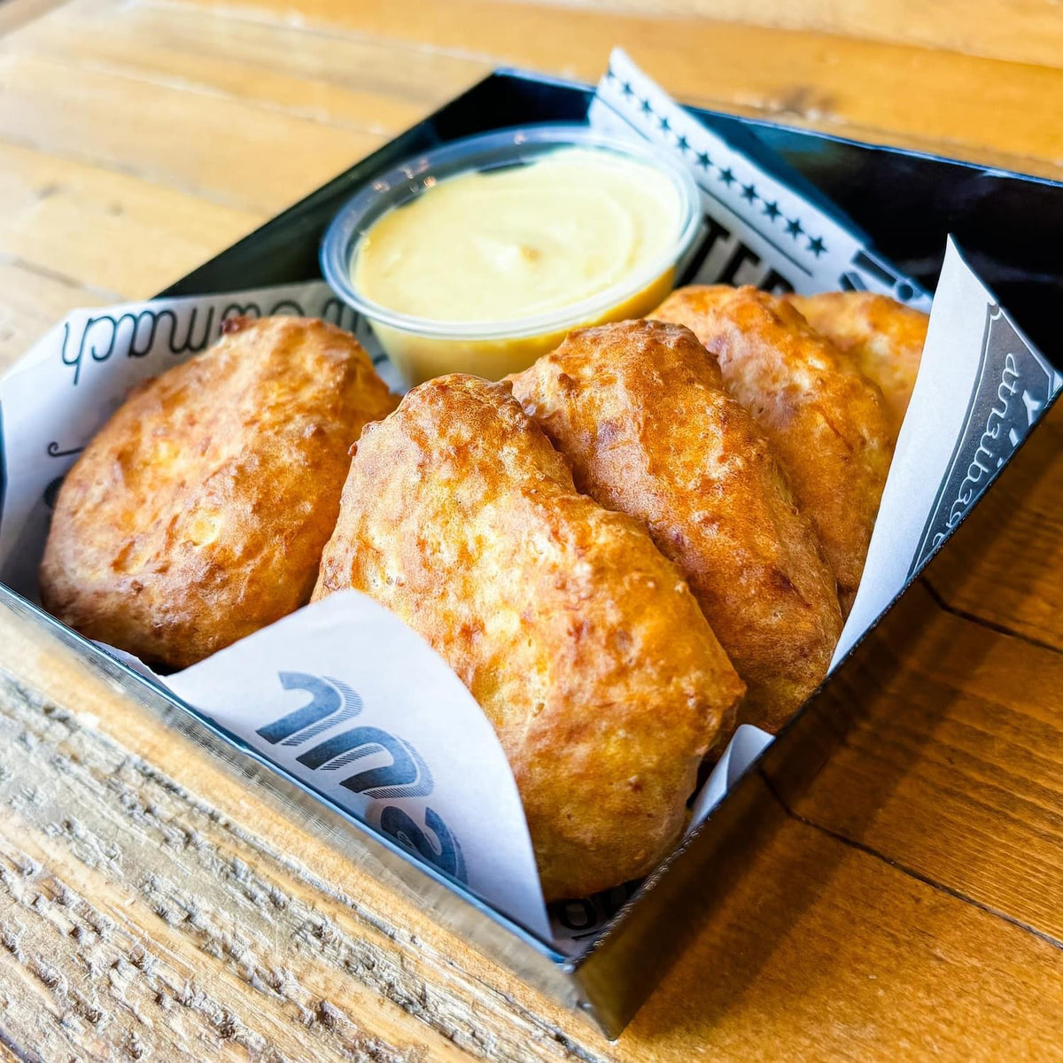 Square black box filled with round, golden brown chicken nuggets with a portion cup of creamy pale yellow dipping sauce.