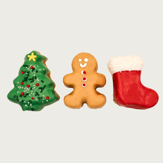 Three Christmas-themed dog cookies shaped like a Christmas tree with ornaments, decorated gingerbread man, and red & white stocking.