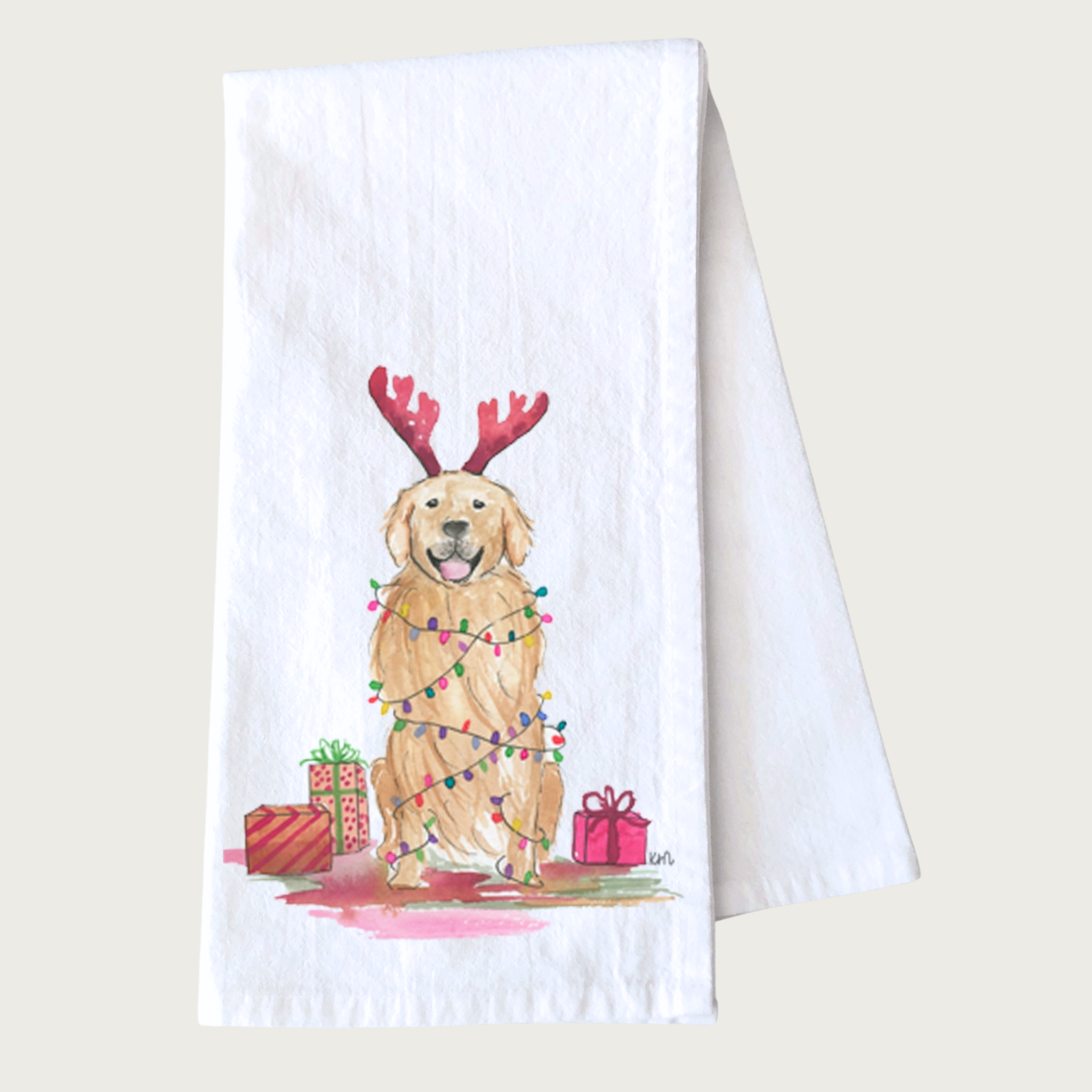 Decorative Christmas-themed tea towel with a Golden Retriever wearing red deer antlers and multicolored Christmas lights draped on its body, and sitting amongst wrapped gifts. White towel with dog illustration that looks like it was done in ink & watercolor.