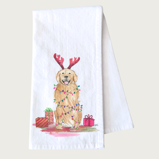 Decorative Christmas-themed tea towel with a Golden Retriever wearing red deer antlers and multicolored Christmas lights draped on its body, and sitting amongst wrapped gifts. White towel with dog illustration that looks like it was done in ink & watercolor.