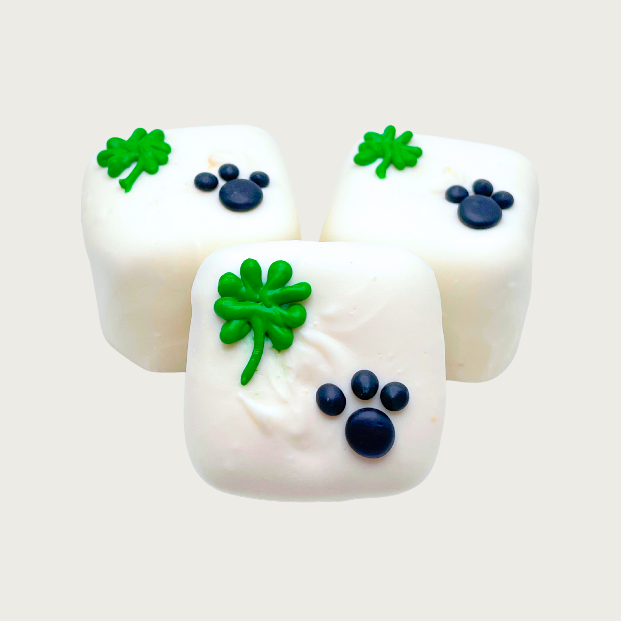 Three square Bon Bon dog treats. They are iced white, and each has a single, iced emerald green four-leafed clover in the top left corner, and the bottom right is a small black paw print.
