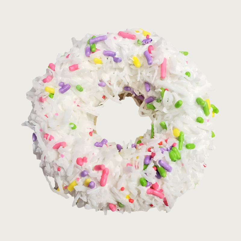 Thick, donut-shaped dog cookie covered with white frosting, shredded coconut, and decorated with pastel sprinkles.