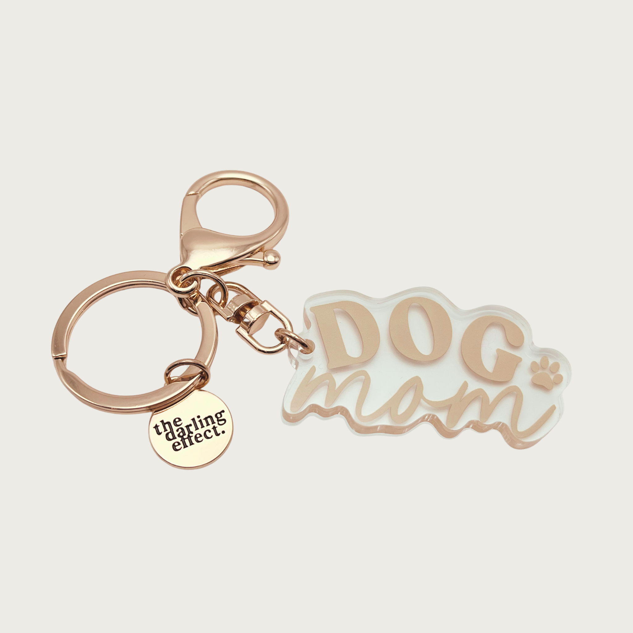 Keychain with 'DOG mom' written in tan on a clear acrylic charm. "DOG" is in all caps, and "mom" is lower case script. There is a small paw print to the right of both words. A standard split keyring and lobster clip are attached to the charm, along with a small gold brand logo charm that reads "the darling effect."