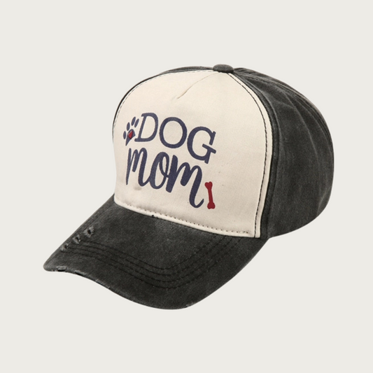 Washed black baseball cap with black 'DOG MOM' text in center (on light beige background), with black & red paw print and red dog bone accent. "Dog" is in all caps serif text, "Mom" is in all caps script text. Rim has 3 small spots of intentional distressing.