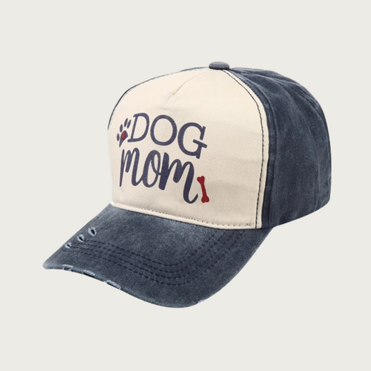 Washed dark navy blue baseball cap with black 'DOG MOM' text in center (on light beige background), with black & red paw print and red dog bone accent. "Dog" is in all caps serif text, "Mom" is in all caps script text. Rim has 3 small spots of intentional distressing.